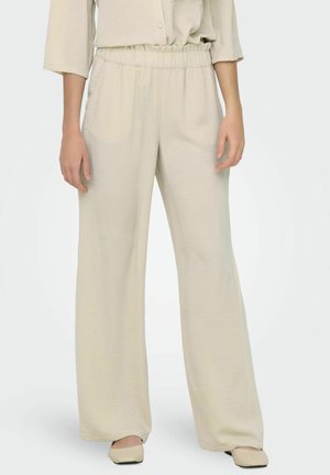 Donna che indossa pantaloni beige larghi con vita elastica e scarpe beige abbinate con punta quadrata, in piedi contro uno sfondo neutro.