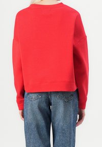 Sweat-shirt rouge court avec épaules tombantes et poignets côtelés, fabriqué en tissu doux. Porté avec un jean bleu taille haute, doté de poches arrière.