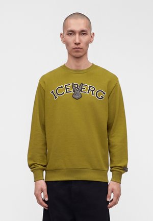 Mężczyzna w oliwkowej bluzie z napisem „ICEBERG” i skrzydlatym emblematem, stojący na jednolitym białym tle.