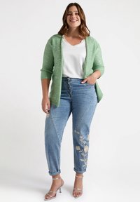 Ljust grönt stickat cardigan med glittrande detaljer, vit V-ringad topp, blå broderade jeans med blommönster och silvriga sandaler med remmar.