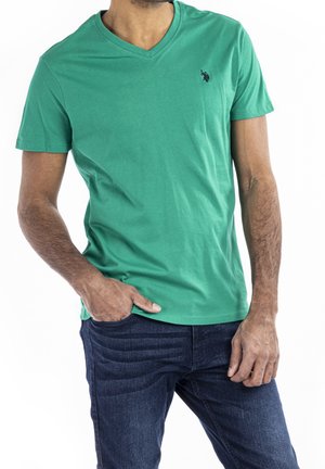Man draagt een groene V-hals T-shirt met een klein donker logo op de borst, ingestopt in een donkerblauwe spijkerbroek, met één hand in de zak.