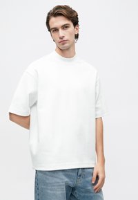 Ung mann med lys hud og brunt hår iført en enkel hvit kortermet skjorte og lyseblå jeans, som står mot en hvit bakgrunn.