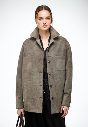 Frau mit hellbraunen Haaren trägt über einem schwarzen Outfit eine übergroße Wildlederjacke in Taupe und hält eine dunkelbraune Ledertasche vor weißem Hintergrund.