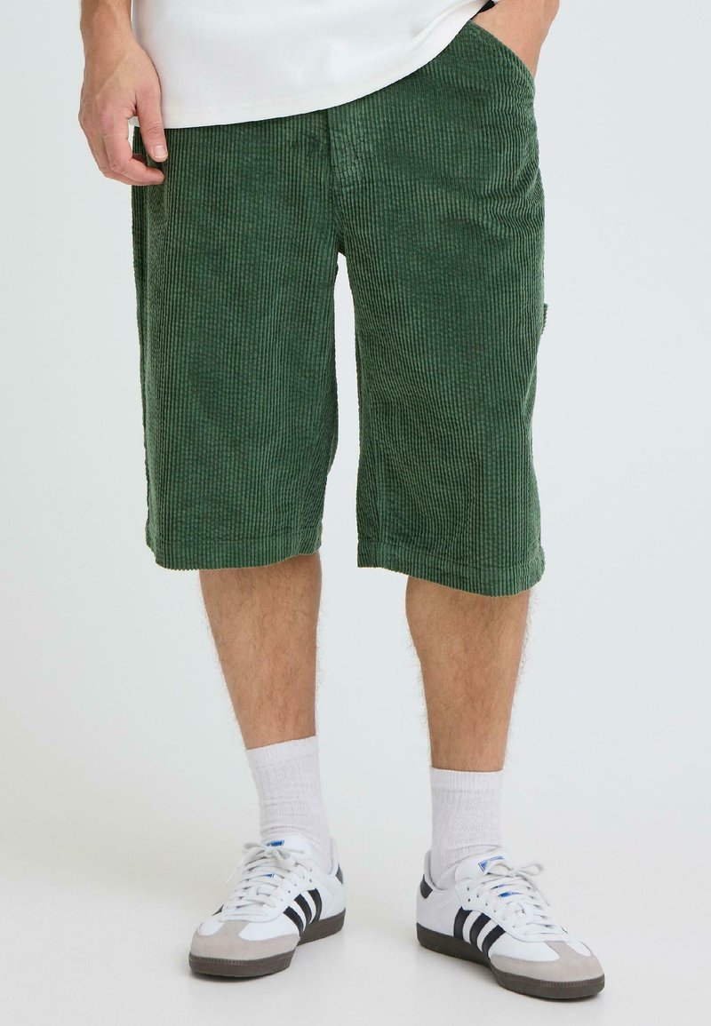 Shorts en velours côtelé vert avec un motif texturé, dotés de poches latérales et d'une coupe mi-genoux, assortis à des chaussettes blanches et des chaussures blanches.