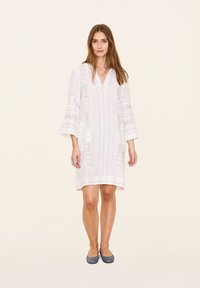 Weißes besticktes Kleid mit Dreiviertelärmeln, V-Ausschnitt und komplizierten Mustern auf dem Stoff. Getragen mit blauen Ballerinas.