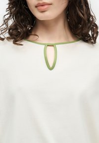 Γυναίκα που φοράει λευκό τοπ με άνοιγμα στο λαιμό που είναι περιμετρικά διακοσμημένο με πράσινο και έχει καστανά σγουρά μαλλιά που φαίνονται στους ώμους.