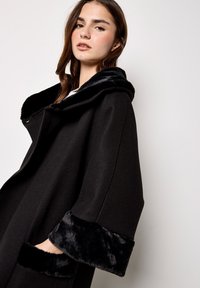 Schwarzer Mantel mit Kapuze, der einen weichen Kunstfellbesatz hat. Lockere Passform mit weiten Ärmeln und Seitentaschen. Glatte Stofftextur.