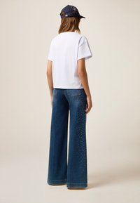 Witte t-shirt met korte mouwen, gecombineerd met blauwe high-waisted wijde jeans met zilveren studs langs de buitenste naden. Marineblauwe pet met versieringen.
