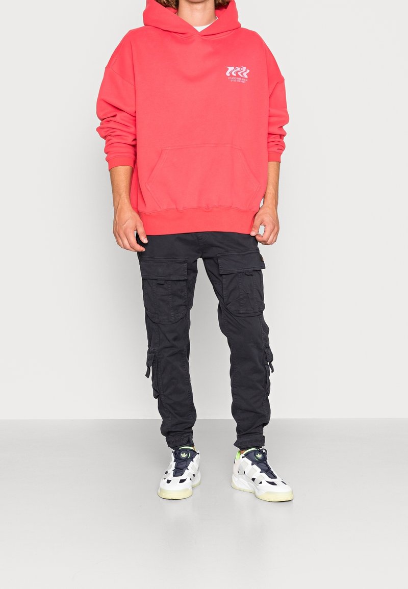 Personne portant un sweat à capuche rose vif, un pantalon cargo noir et des baskets blanches avec des touches foncées, debout devant un fond clair et uni.