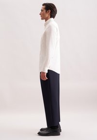 Man in wit knoopshirt, marineblauwe broek en zwarte schoenen zijwaarts staand op een effen lichte achtergrond.