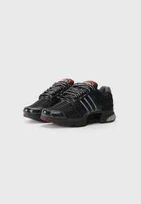 Chaussures de sport noires avec une tige en mesh, présentant des accents à rayures blanches, une semelle texturée et des détails rouges sur le talon et la languette.