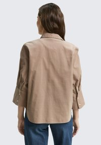 Bruine kraagshirt met lange mouwen, knoopmanchetten en een licht gebogen zoom. De stof lijkt soepel met een minimale textuur.
