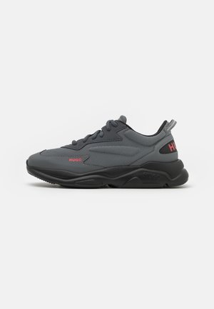 Zapatilla deportiva de color gris oscuro con suela negra, cordones negros y la marca "HUGO" en rojo en el lateral y la lengüeta del talón.