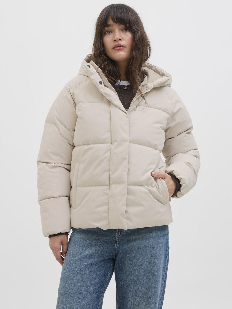 Beige puff jacket med hætte, der har en quiltet overflade, frontlommer og lynlås lukning. Båret over en mørk sweater og blå jeans.