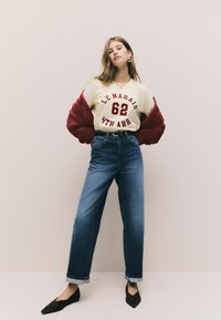 Tricou crem cu imprimeu roșu, pulover din blană burgundă, blugi cu talie înaltă de culoare albastră și pantofi negri cu vârf ascuțit, având un model subtil.
