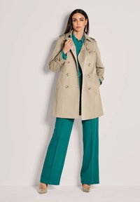 Vrouw in een turquoise shirt en broek, gekleed in een beige trenchcoat, poseert met één hand in de zak en de andere hand die de kraag van de coat vasthoudt.