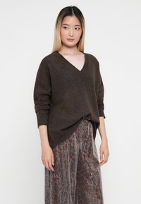 Bruine wollen trui met een V-hals, drop shoulders en een relaxed fit, gecombineerd met een gestreepte plissémidi-jurk in grijstinten en rood.