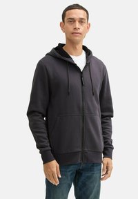 Felpa con zip di colore grigio scuro, dotata di tasca frontale, cappuccio con cordino regolabile e texture di tessuto combinato, indossata sopra una maglietta bianca.