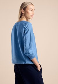 Blusa azzurro chiaro, con texture, dalla vestibilità comoda e maniche a 3/4. Presenta un collo rotondo e polsini elasticizzati. Abbinata a pantaloni blu scuro.
