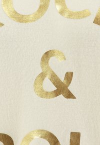 Symbole esperluette métallique doré centré sur un fond en tissu écru texturé avec des lettres dorées partielles aux bords supérieur et inférieur.