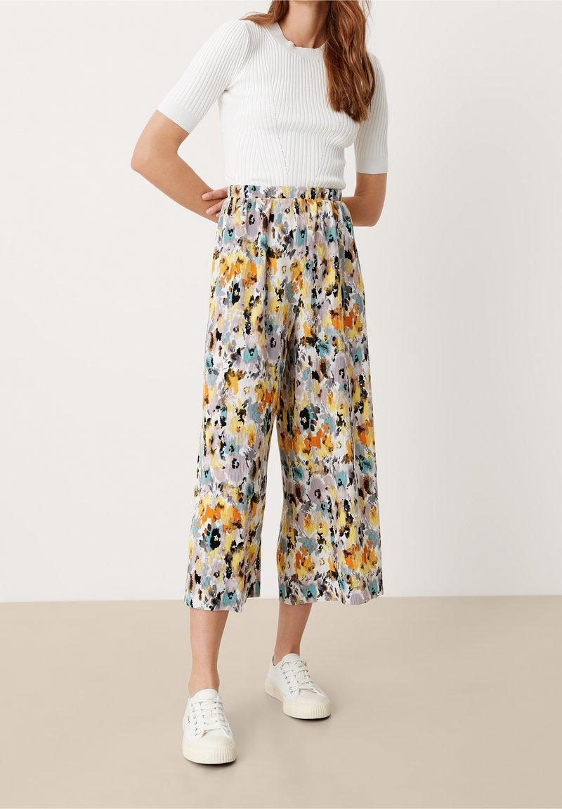 Femme portant un haut blanc côtelé à manches courtes, un pantalon large fleuri coloré et des baskets blanches, debout sur un sol beige contre un mur uni.