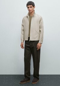 Veste légère beigne avec col montant, fermeture éclair et poches latérales, associée à un pantalon marron foncé et des chaussures marron. Texture lisse.