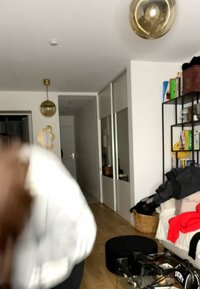 Osoba pochylająca się do przodu w białym swetrze w salonie z szklanym stolikiem kawowym, regałami na książki i wiszącymi kulistymi lampami.