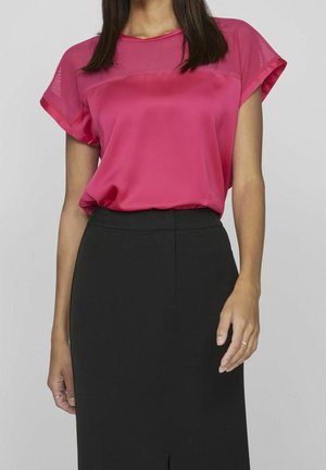 Femme portant un chemisier rose vif à manches courtes rentré dans une jupe noire taille haute arrivant au genou, sur un fond clair uni.