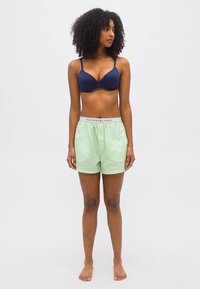 Γυναίκα με σγουρά μαλλιά που φοράει μπλε σουτιέν και ανοιχτό πράσινο σορτς Polo Ralph Lauren, όρθια, ξυπόλητη, μπροστά σε λευκό απλό φόντο.