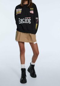 Sudadera negra con parches de temática de carreras, combinada con una falda plisada beige. Botas de plataforma negras con cordones y calcetines tobilleros blancos completan el look.