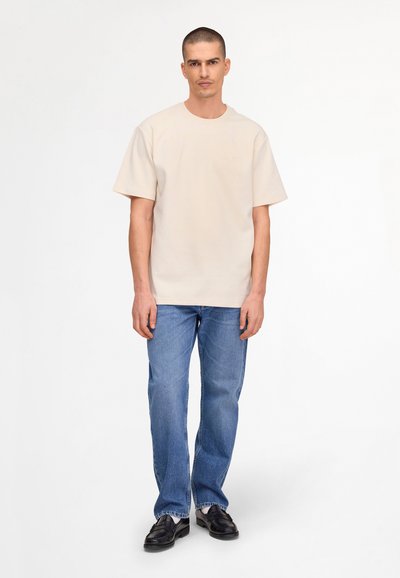 Mann steht und trägt ein beigefarbenes Kurzarm-T-Shirt, blaue Jeans und schwarze Loafers vor weißem Hintergrund.