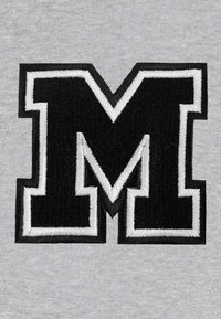 Lettera "M" ricamata in nero, contornata in bianco, posizionata su uno sfondo di tessuto grigio texturizzato. Il design presenta angoli affilati e un chiaro contrasto.