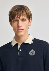 Marineblå polo med en kremfarvet krave, der har et logo med en krone og laurbærdesign, samt knaplukning med to knapper. Tekstureret stof med striber.