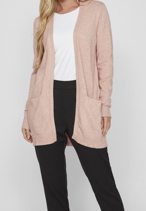 Femme portant un cardigan rose clair ouvert avec des poches, par-dessus un haut blanc et un pantalon noir, devant un fond gris.
