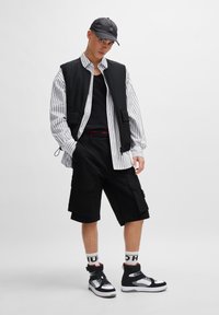 Black vest over a white and black striped shirt, black shorts with pockets, and black high-top sneakers. Accented with branded white socks.