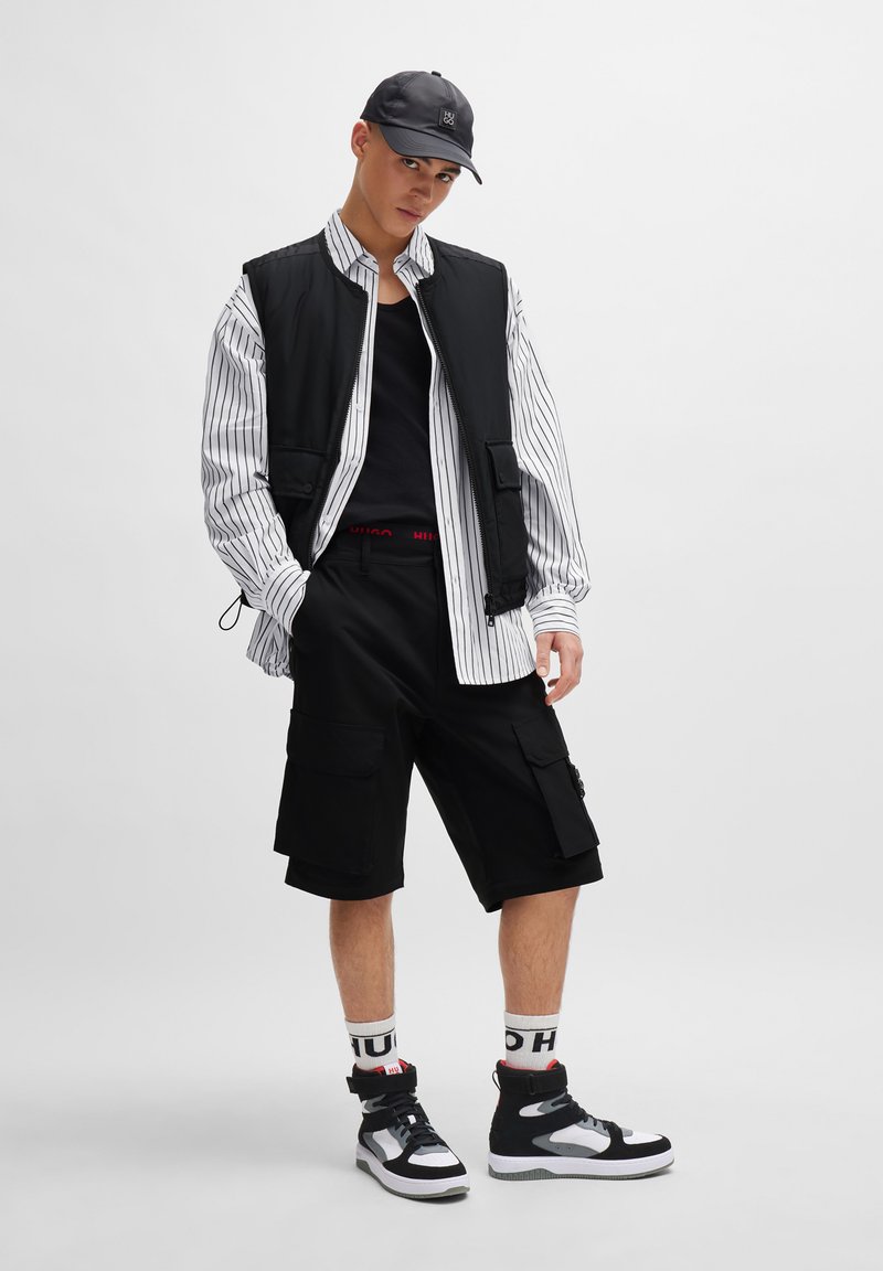 Black vest over a white and black striped shirt, black shorts with pockets, and black high-top sneakers. Accented with branded white socks.