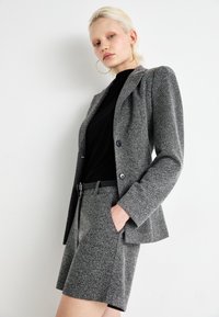 Blazer gris texturé avec épaules rembourrées et deux boutons, associé à un short assorti, porté par-dessus un haut noir.