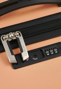 Chiusura con cerniera a due tiranti in metallo argento, una traccia di cerniera nera testurizzata e un lucchetto approvato dalla TSA su uno sfondo color corallo.