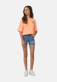 Heldere oranje oversized t-shirt met zwart logo, gecombineerd met blauwe denim shorts. Model draagt witte sneakers. Eenvoudige, casual outfit.