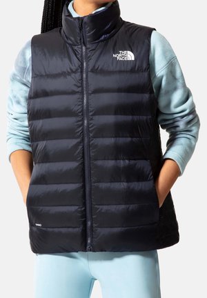 Personne portant une doudoune noire avec le logo The North Face sur une chemise bleu clair à manches longues et un pantalon bleu clair assorti.