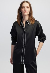 Camicia in satin nera con maniche lunghe, dotata di chiusura a bottoni e cuciture bianche a contrasto lungo il colletto e la patta.