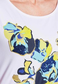 Witte T-shirt met multicolore bloemapplicaties met blauwe, gele en zwarte details; versierd met pailletten en gouden accenten.