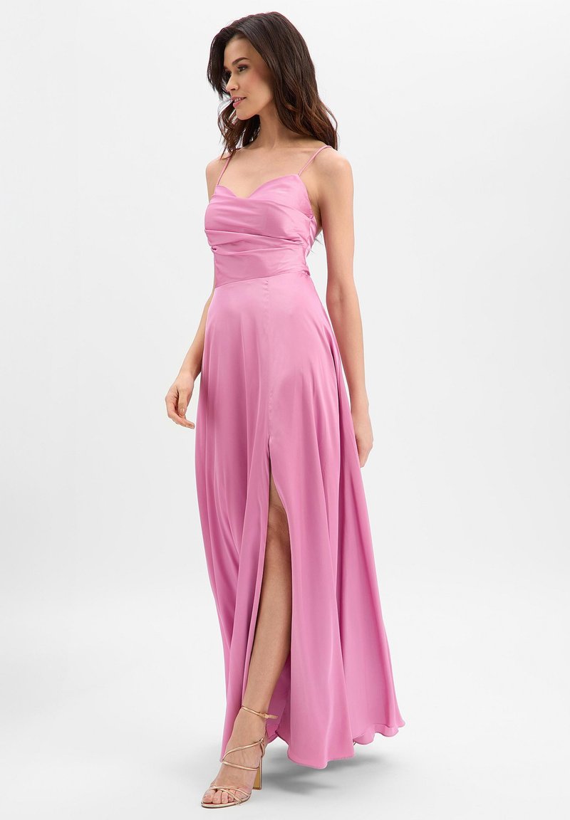 Femme portant une robe longue en satin rose avec des bretelles spaghetti et une fente haute sur la cuisse à l'avant, associée à des talons ouverts dorés.