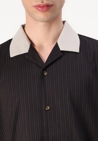Camicia a righe nere con colletto beige, dotata di chiusura a bottoni e tessuto testurizzato. Il design include righe verticali.