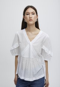Blouse en coton blanc avec un col en V, des manches courtes à volants et une taille froncée. Présente des petits pois texturés subtils sur le tissu.