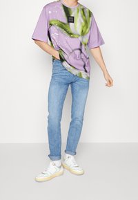 Lila oversized t-shirt med grön abstrakt design och svart logotyp-patch, kombinerad med ljusblå jeans och flerfärgade sneakers.