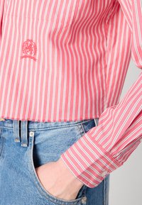 Röd och vit randig skjorta med en logotyp på vänster bröst, långärmad och med knäppning vid ärmsluten. I kombination med ljusblå denimjeans.