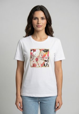 Donna con capelli lunghi e mossi che indossa una t-shirt bianca con una stampa quadrata floreale colorata, abbinata a jeans azzurri, su uno sfondo grigio.