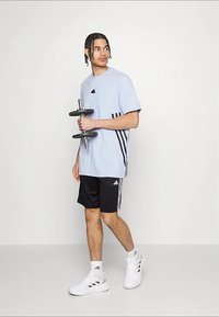 Jauns vīrietis tur hanteli, valkājot gaiši zilu T-kreklu un melnas šortus ar baltām sporta kurpēm, stāvot pret vienkrāsainu baltu fonu.