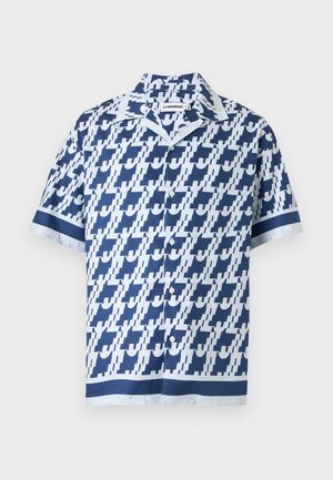 Homme debout les mains dans les poches portant une chemise à manches courtes bleue et blanche à motifs, un pantalon blanc et des baskets bleu marine sur fond blanc.
